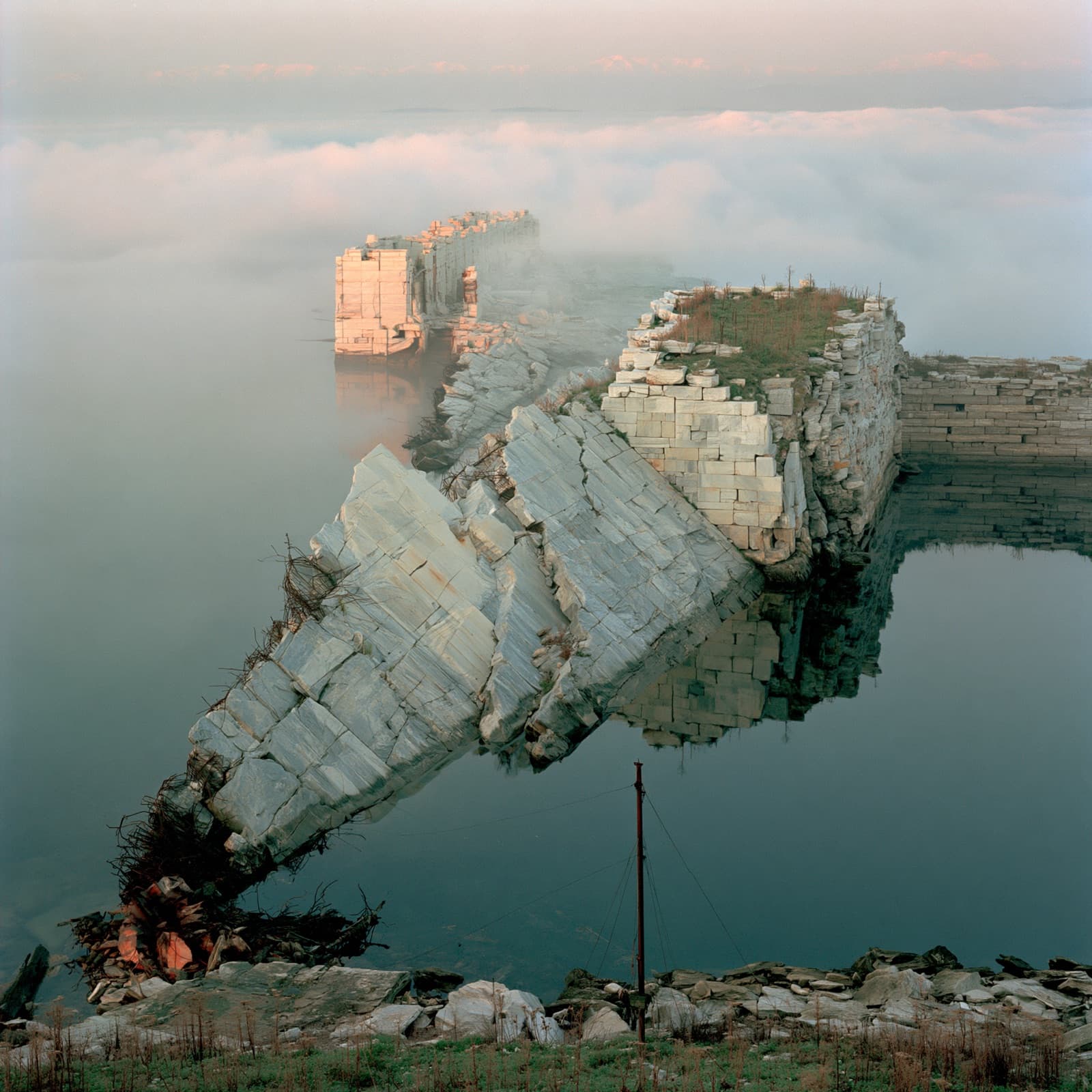 Quarried marble blocks still standing in still water at dawn, fog beyond them.